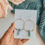 A hand holds a card with JaxKelly's Amazonite Hoop - Hope gold-plated earrings, featuring green amazonite stone pendants. The card says "Wear Good Vibes" and "Amazonite hope stone," set against pink flowers and blue fabric.