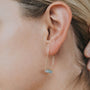 Close-up of a person’s ear wearing the Amazonite Gemstone Drop - Hope earring, featuring a rectangular light blue stone and elegant 18k gold plating. Blonde hair is pulled back to showcase the minimalist jewelry.