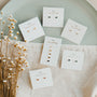 Six pairs of Lilac Baguette minimalist stud earrings with sterling silver posts rest on white display cards atop a pale green plate, accompanied by small white flowers and a beige textured cloth.