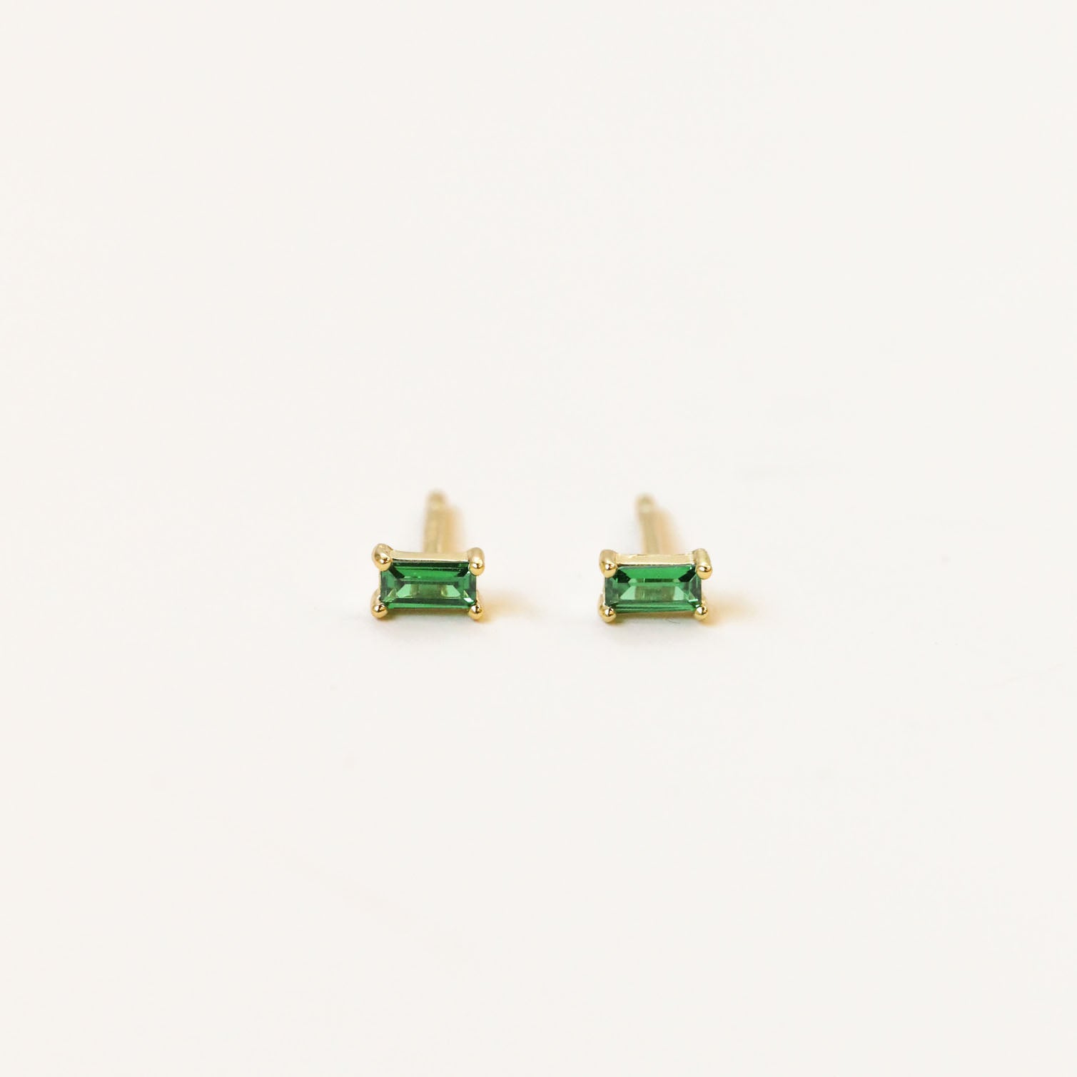 The Emerald Baguette features gold-plated stud earrings, each set with a small rectangular green gemstone, shown against a plain white background.
