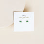 A pair of small, rectangular green Emerald Baguette stud earrings are displayed on a white card with gold "JaxKelly wear good vibes" text, set against a beige background.