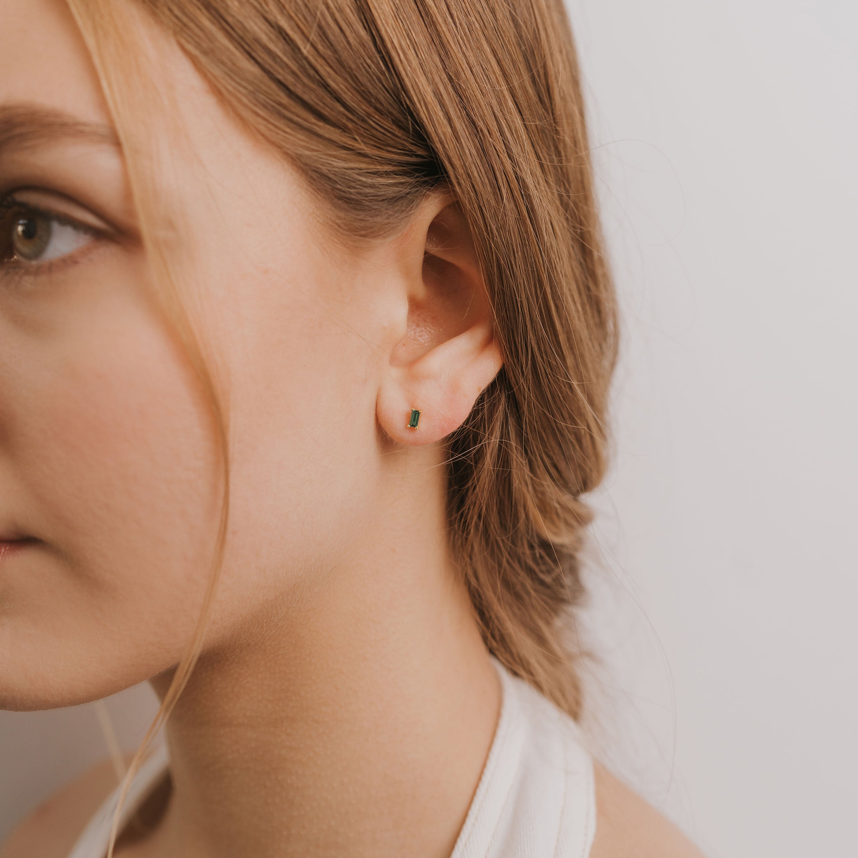 Close-up of a young woman with light brown hair wearing the Emerald Baguette—a small green sterling silver earring—in her left ear, with loose strands framing her face against a plain white background.