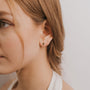 Close-up of a young woman with light brown hair wearing the Emerald Baguette—a small green sterling silver earring—in her left ear, with loose strands framing her face against a plain white background.