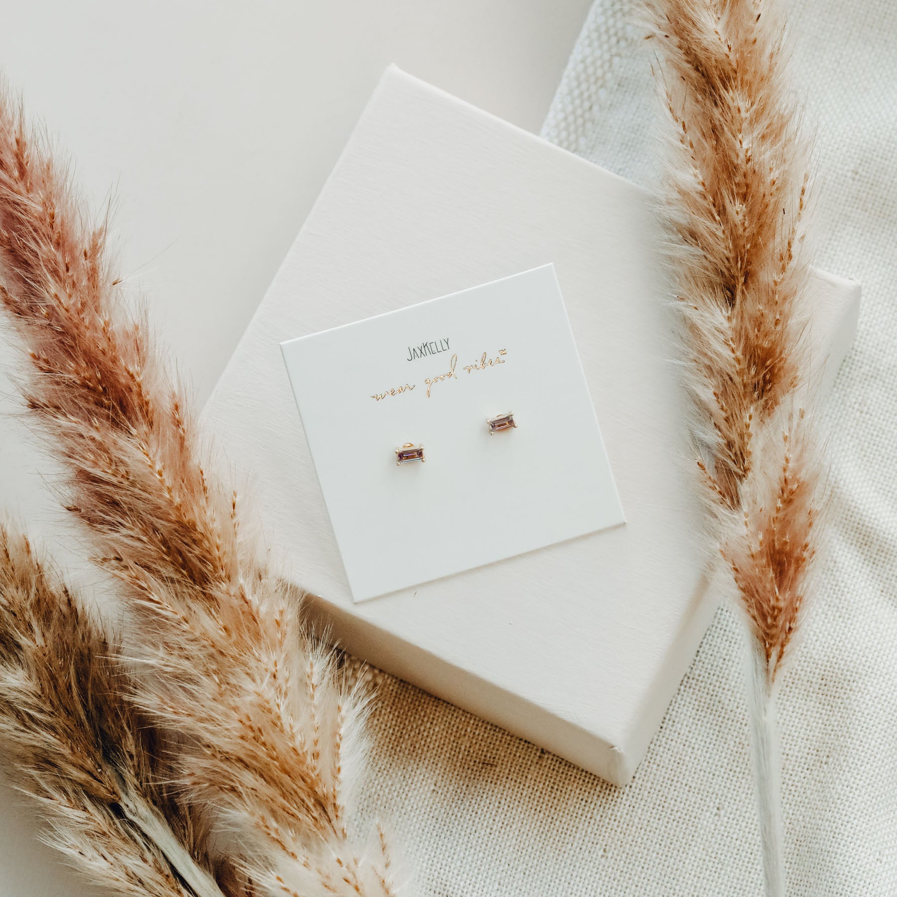 The Lilac Baguette minimalist stud earrings with sterling silver posts are displayed on a white card on a box, surrounded by beige pampas grass and textured fabric.