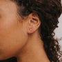 Close-up of a person’s ear wearing the Lilac Baguette, a small rectangular stud earring with 18k gold plating. The wearer has medium brown skin and curly dark hair, with a plain neutral background.