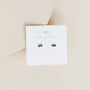 A pair of Sapphire Baguette small rectangular blue stud earrings with gold settings are displayed on a white card reading "JAY KELLY wear good vibes" in gold, resting on a neutral beige surface.
