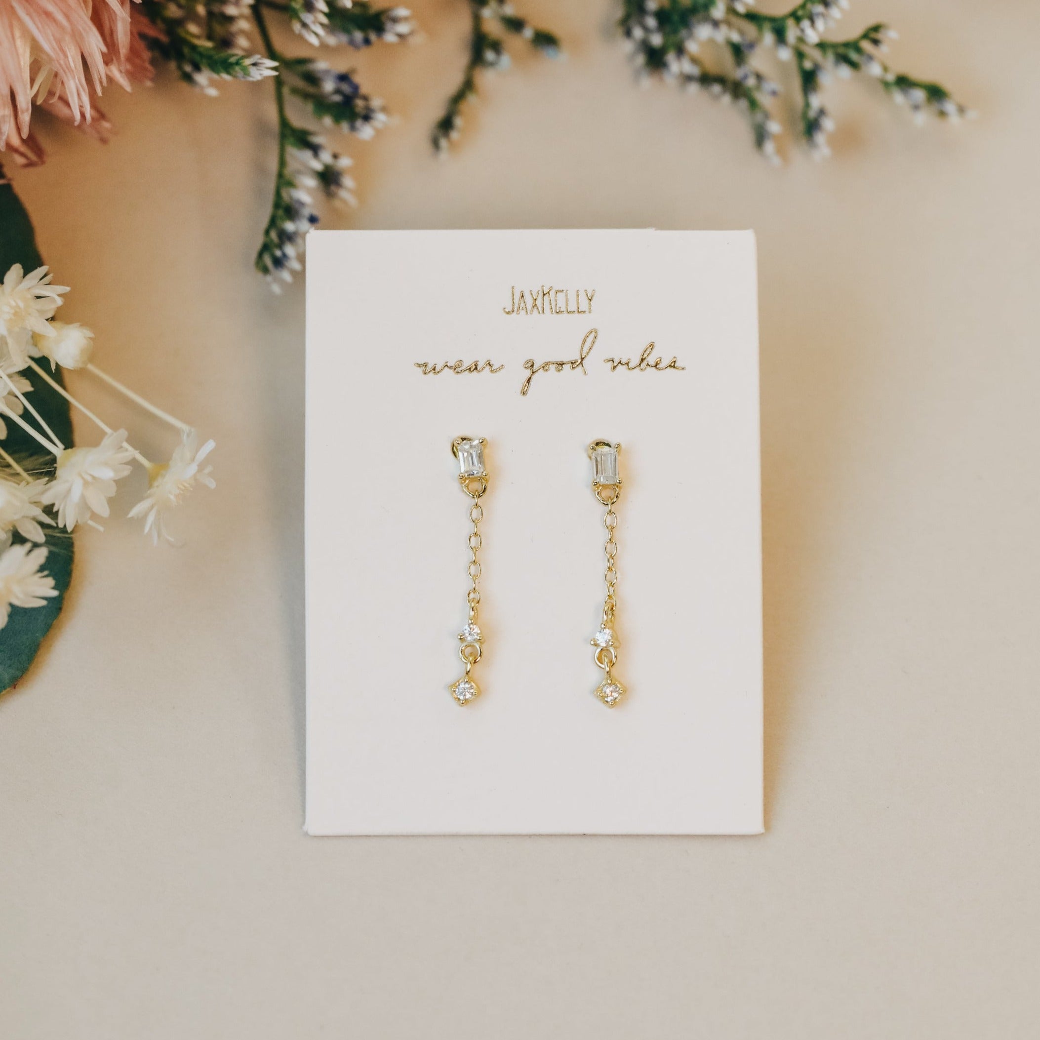 The Baguette Drop earrings, featuring 18k gold plating and small crystals, are shown on a white card that says "JaxKelly wear good vibes" and are surrounded by delicate flowers.