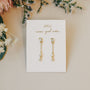 The Baguette Drop earrings, featuring 18k gold plating and small crystals, are shown on a white card that says "JaxKelly wear good vibes" and are surrounded by delicate flowers.