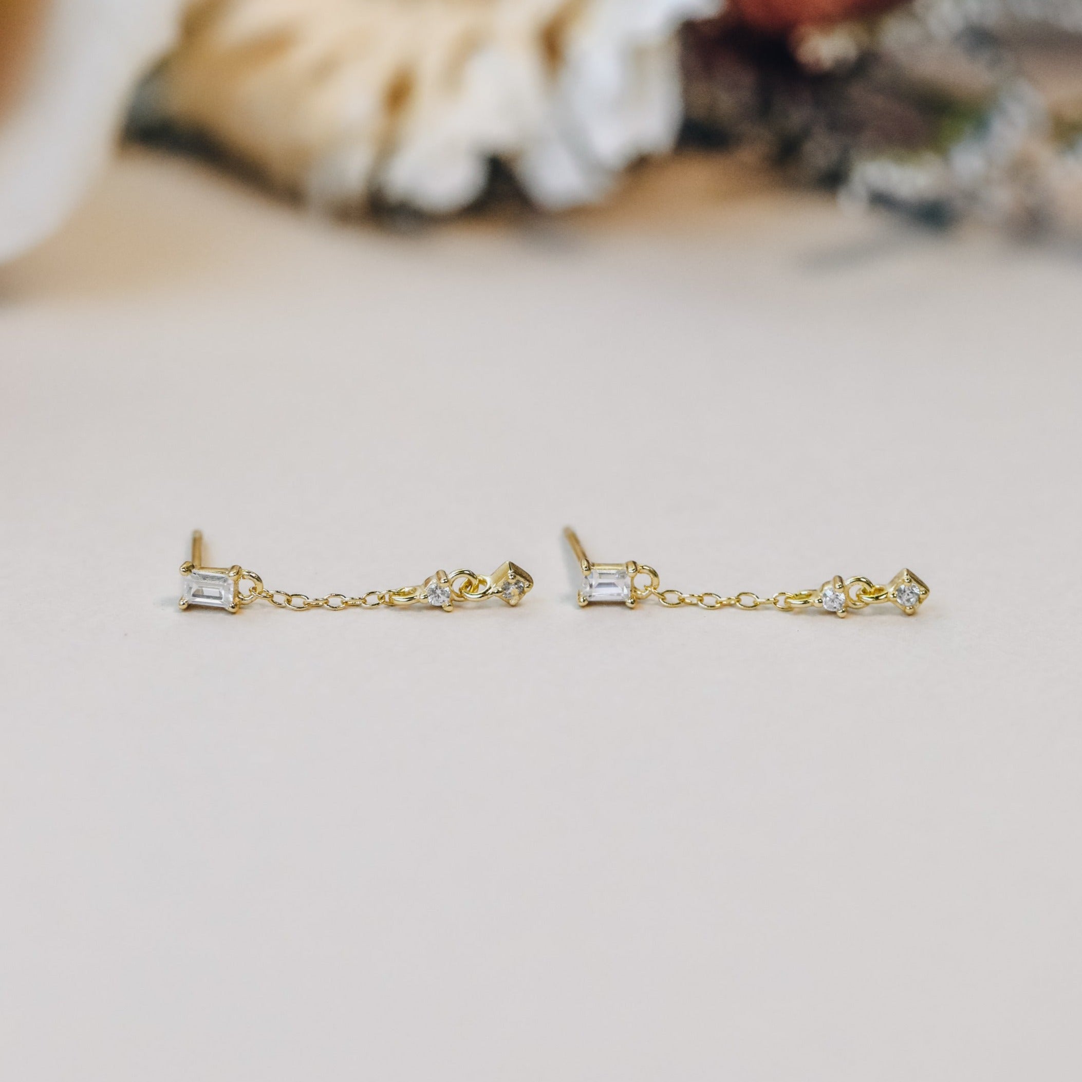 The Baguette Drop features delicate 18k gold plated dangle earrings with small rectangular and diamond-shaped stones, shown on a light surface with a softly blurred background.