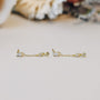 The Baguette Drop features delicate 18k gold plated dangle earrings with small rectangular and diamond-shaped stones, shown on a light surface with a softly blurred background.