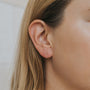 Close-up of a person with light skin and straight, light brown hair tucked behind the ear, wearing the Minimalist Bar earring—18k gold plated for a refined touch. The background is softly blurred for emphasis.