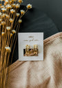 The Bold Hoop, a pair of 18k gold plated hoop earrings, sits on a white JaxKelly card beside small yellow and white flowers and a textured beige cloth atop a dark round surface.