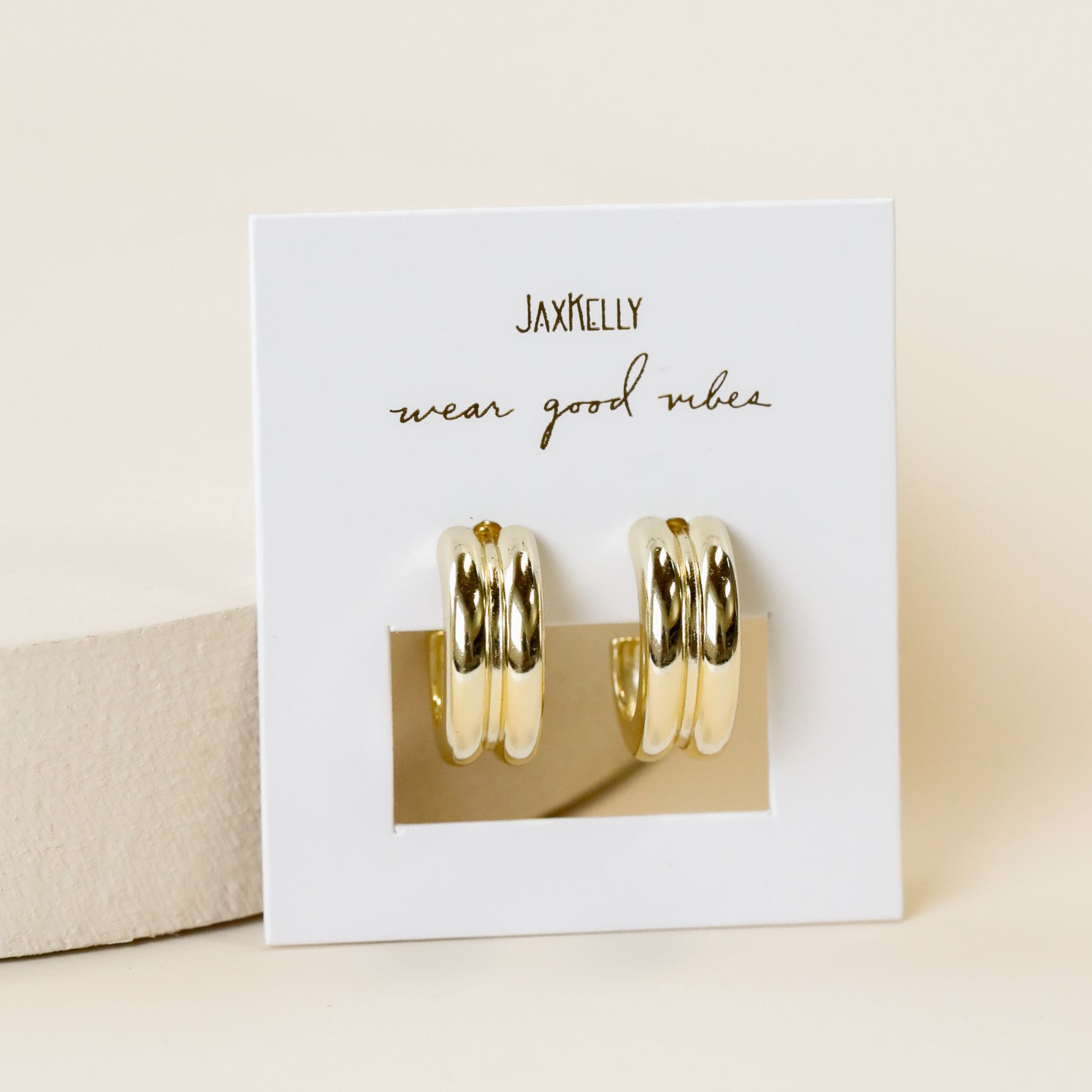 A pair of Bold Hoop 18k gold plated chunky earrings are displayed on a white JaxKelly card reading "wear good vibes" in cursive, set against a soft beige background.
