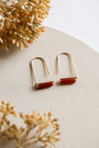 The Carnelian Agate Gemstone Drop - Creativity earrings, featuring minimalist gold plating and rectangular red stones, are displayed on a light textured surface surrounded by dried beige flowers.