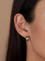 Close-up of a woman's ear wearing the Bold Hoop, a small, shiny 18k gold plated earring. Her dark brown hair and cheek are partially visible against a beige background.