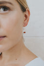 Close-up of a woman with light skin and blonde hair wearing a white top, showcasing her ear adorned with the Carnelian Agate Gemstone Drop - Creativity gold plated earring featuring a small brown stone.