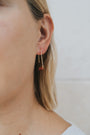 Close-up of a person wearing the Carnelian Agate Gemstone Drop - Creativity earring, featuring a gold-plated drop with a small reddish-brown stone. Blonde hair is tucked behind the ear against a softly blurred neutral background.