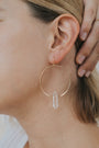 Close-up of a woman's ear wearing the Clear Quartz Hoop - Healing earring, featuring a large handcrafted gold hoop with a clear quartz crystal pendant; her hand touches her hair near the ear.