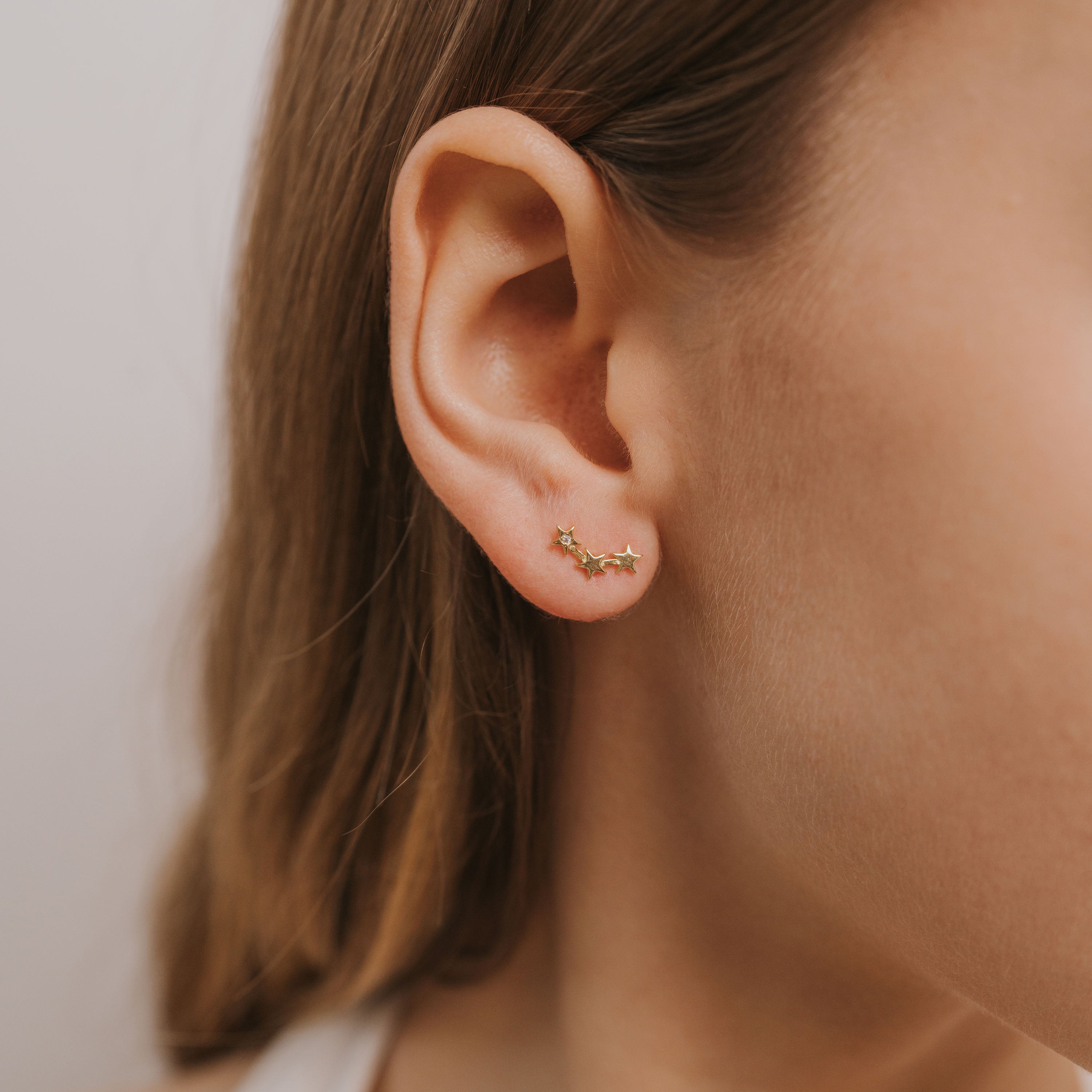 Close-up of a woman’s ear with light brown hair, wearing the Star & Constellation earring—an elegant 18k gold-plated piece shaped like a curved branch, adorned with tiny stones for a delicate celestial look.