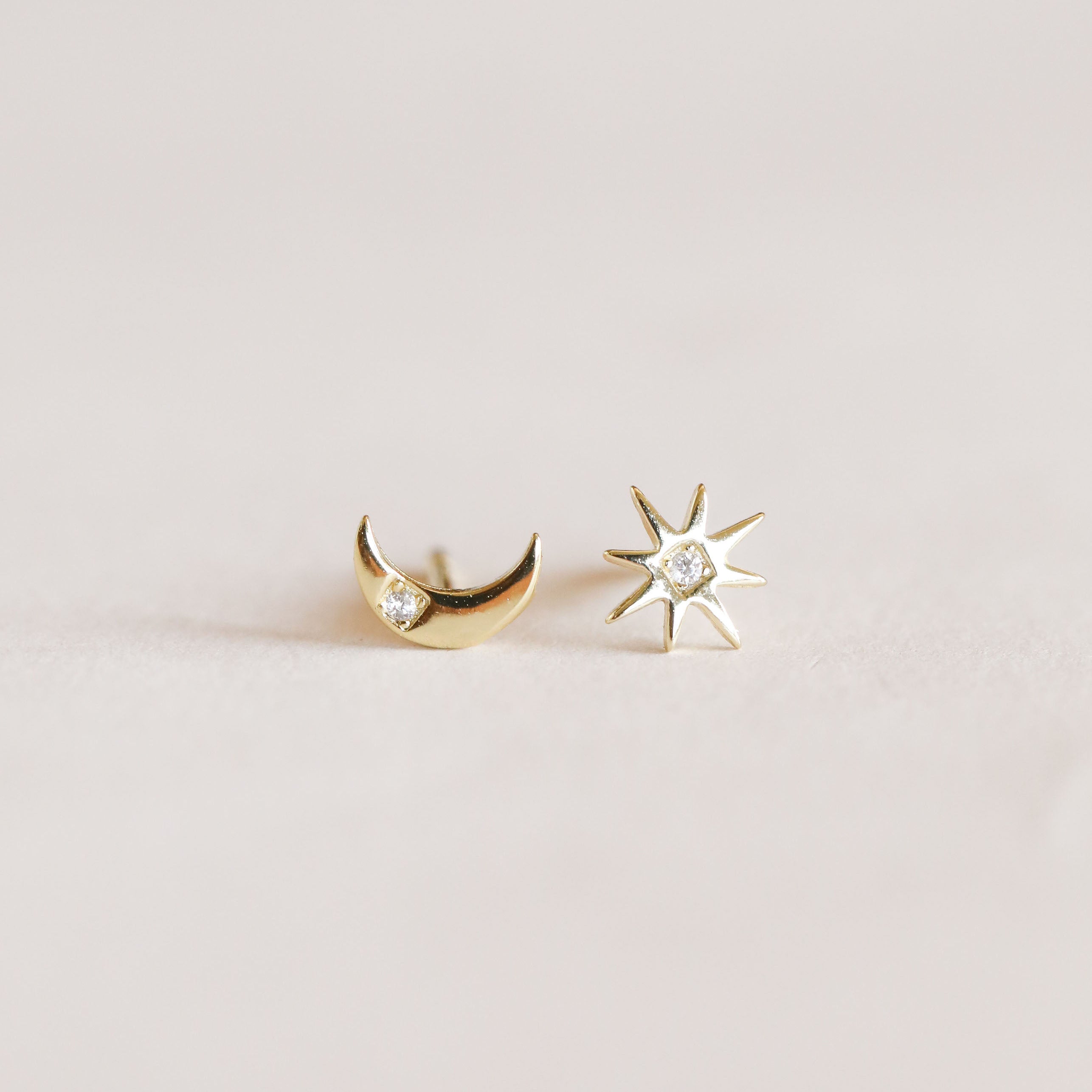 The Sun & Moon features mismatched 18k gold plated stud earrings: one crescent moon with a gemstone and one eight-pointed star with a center stone, displayed on a light background. Perfectly pairs with our Sun & Moon necklace.