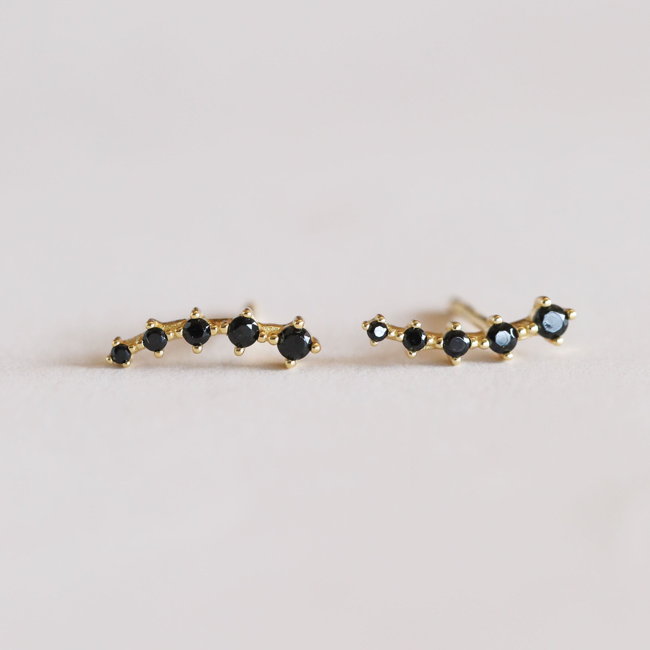 The Black Crawler is a pair of gold earrings with a row of small, round black gemstones set in a curved line, crafted with 18k gold plating and sterling silver posts, shown on a light neutral background.