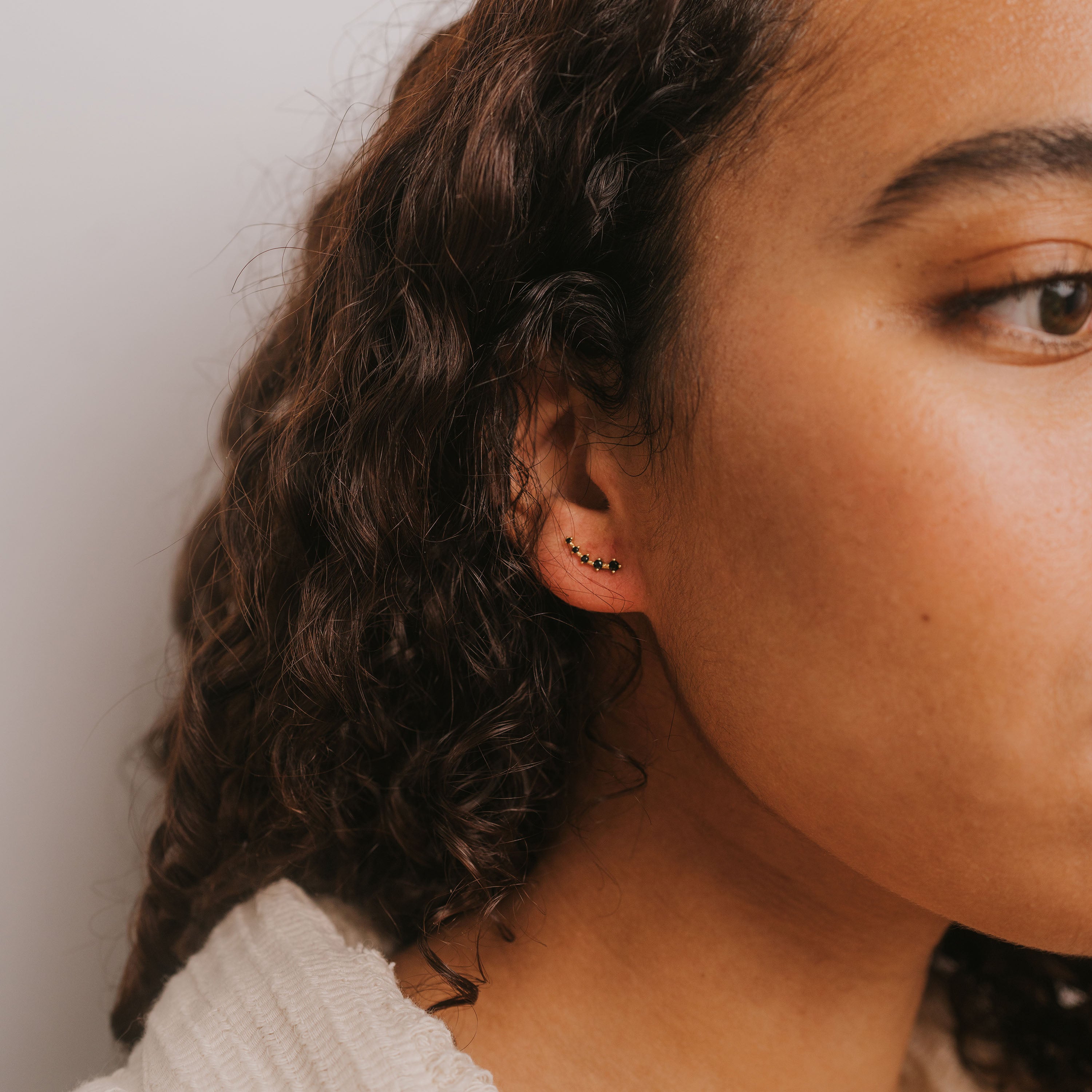 Close-up of a person with curly brown hair wearing the Black Crawler, an ear climber earring shaped like small stars, crafted with 18k gold plating and sterling silver posts. The backdrop is neutral, and they're in a light-textured top.