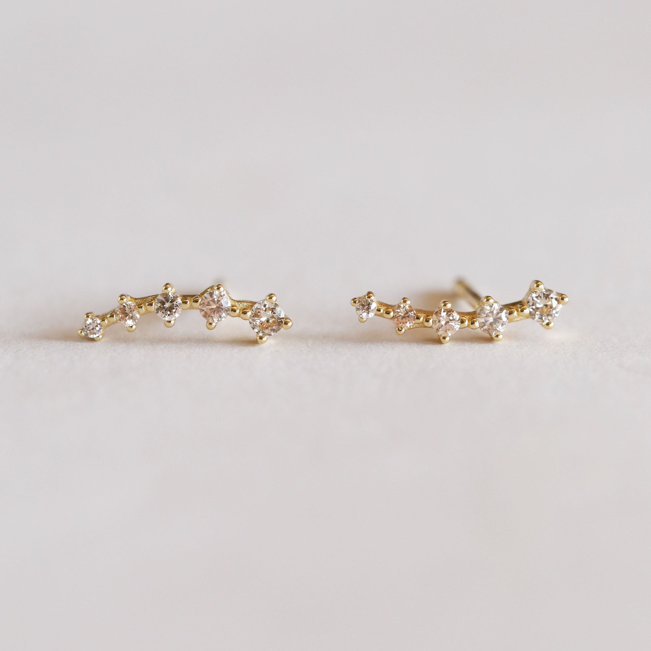 The Champagne Crawler features delicate gold stud earrings shaped like constellations, each adorned with petite sparkling diamonds arranged in a curved line on a soft, light background.