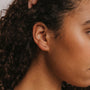 Close-up of a woman's ear with curly hair wearing the Champagne Crawler, a delicate gold ear cuff with small stones and 18k gold plating. The focus is on the crafted, long-lasting jewelry set against a neutral background.