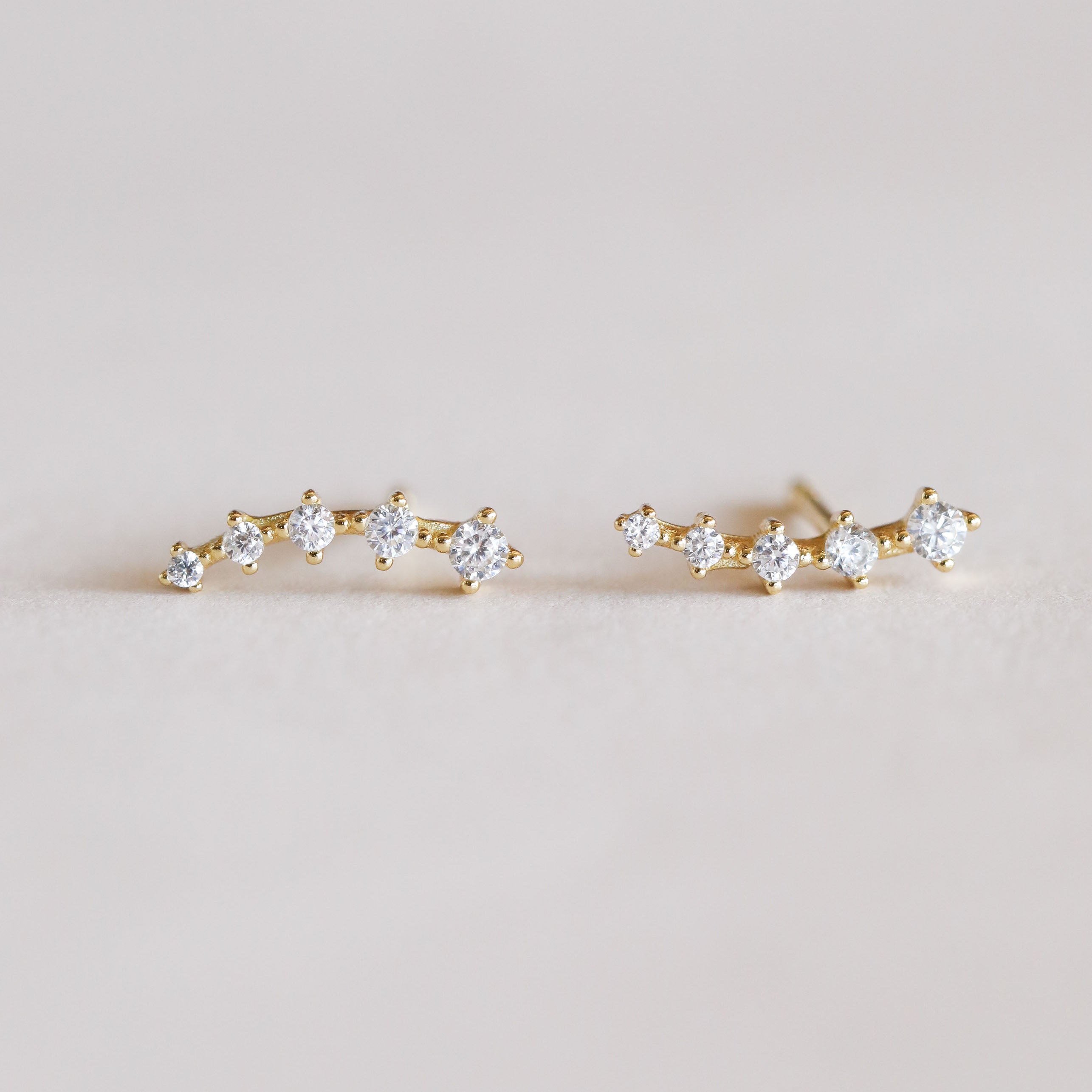 The White Crawler earrings are delicate 18k gold-plated pieces with a curved line of sparkling cubic zirconia, showcased on a neutral background.