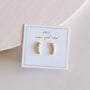 A pair of White Crawler 18k gold-plated cubic zirconia earrings sparkles on a white card labeled “JaxKelly” and “wear good vibes” in gold lettering, set against a pale, textured background.