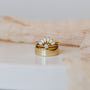 The Opal Crown Ring, featuring a fan arrangement of small white stones, is displayed with other stacking rings—including an 18k gold plated ring with a round diamond—on a white surface and beige textured cloth background.