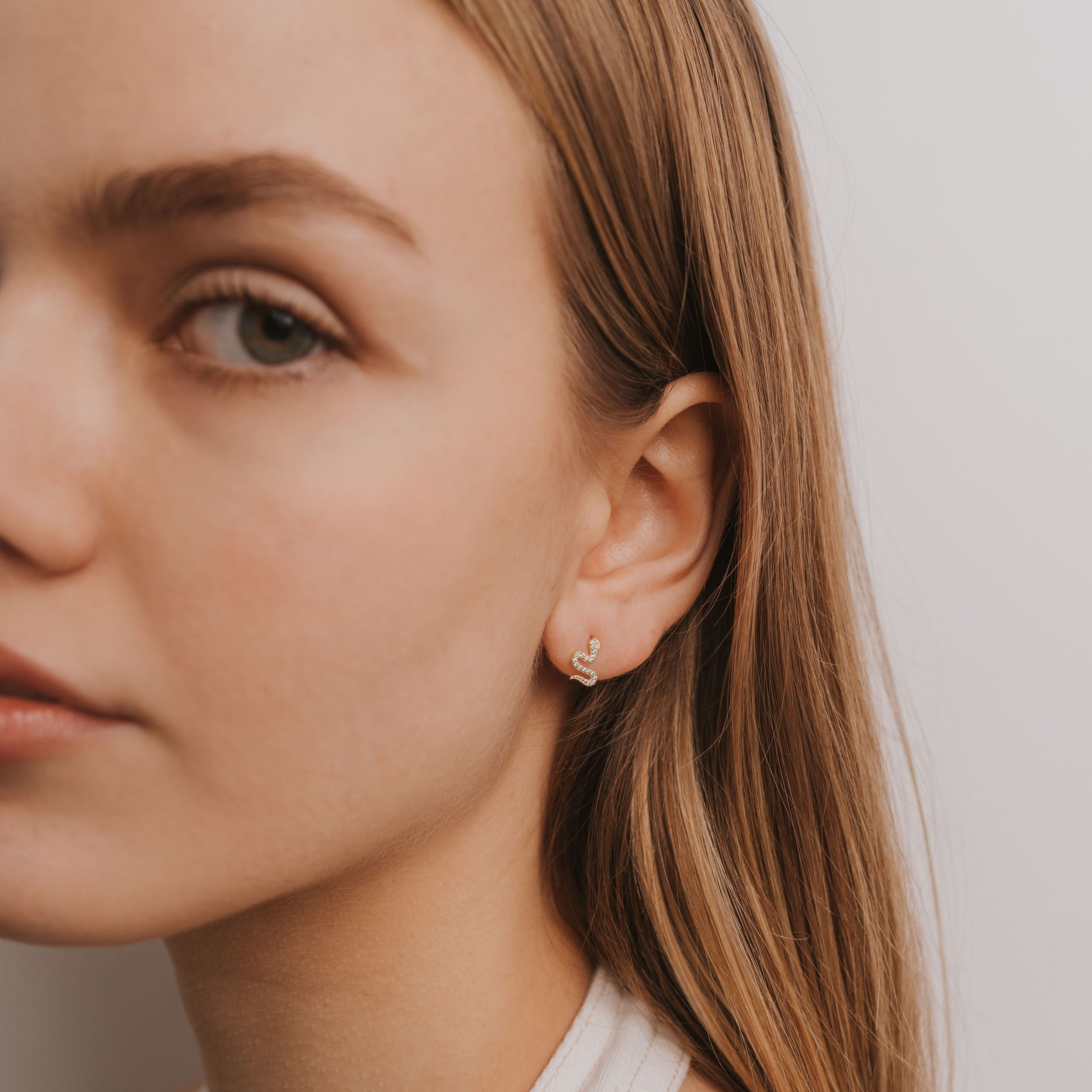 Close-up of a young woman with straight, light brown hair wearing the gold plated Pavé Snake earring—a small, hoop-shaped piece—in her left ear. It glimmers subtly as she faces slightly left with a neutral expression.
