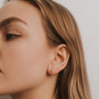 A close-up of a woman’s profile highlights her ear adorned with Baguette Drop 18k gold plated dangle earrings, each featuring a star-shaped charm. She has light skin, straight light brown hair, and the background is plain white.