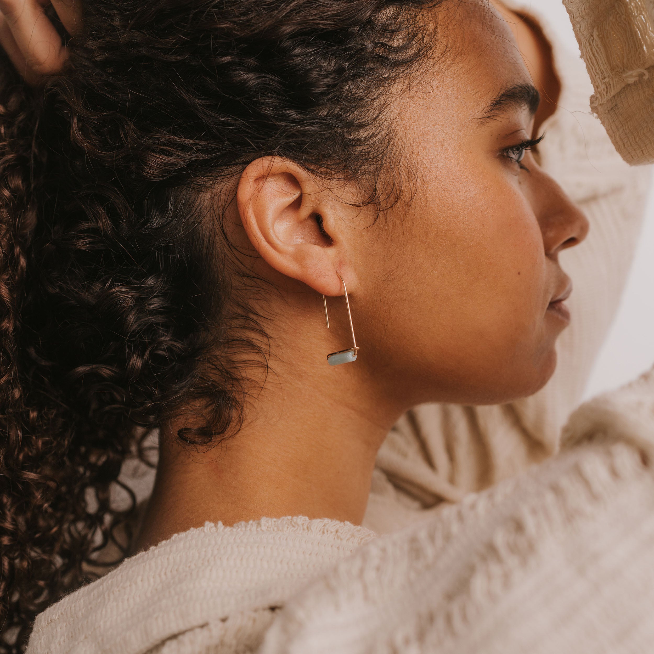 A woman with curly hair wears a textured sweater and the Amazonite Gemstone Drop - Hope earring in 18k gold plate, shown in profile with her arm raised and her gaze forward.