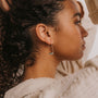 A woman with curly hair wears a textured sweater and the Amazonite Gemstone Drop - Hope earring in 18k gold plate, shown in profile with her arm raised and her gaze forward.