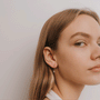 A woman with long, light brown hair and clear skin gazes at the camera, wearing delicate Baguette Drop 18k gold plated dangle earrings and a white top against a soft, neutral background.