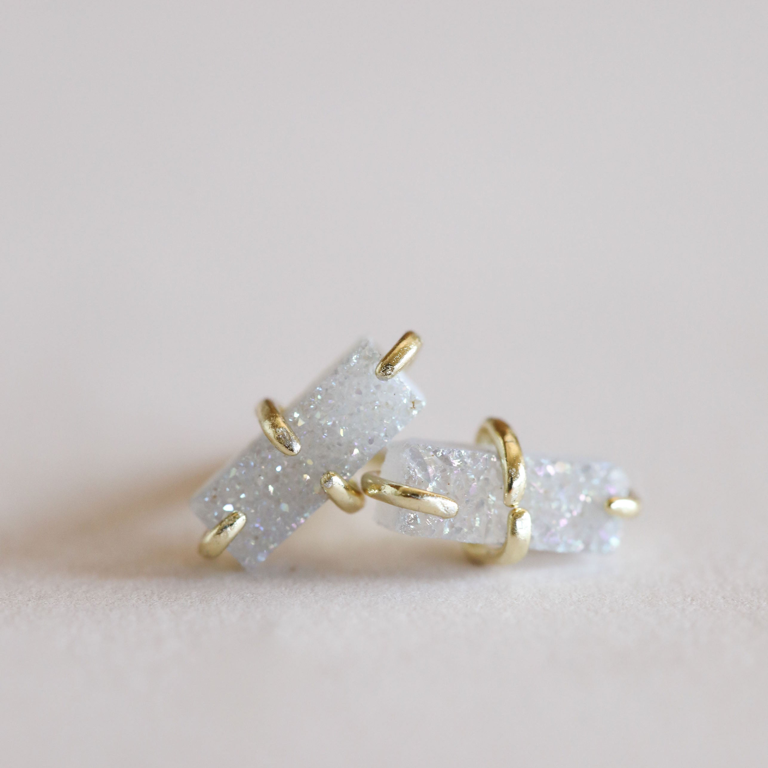 The White Bar Druzy features two small, rectangular white druzy stones set in delicate 18k gold plated prongs and displayed on a soft, pale surface.