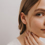 A close-up of a woman with light brown hair tucked behind her ear, wearing White Bar Druzy sterling silver earrings and a white top. Her manicured hand rests on her neck as she displays a calm expression.