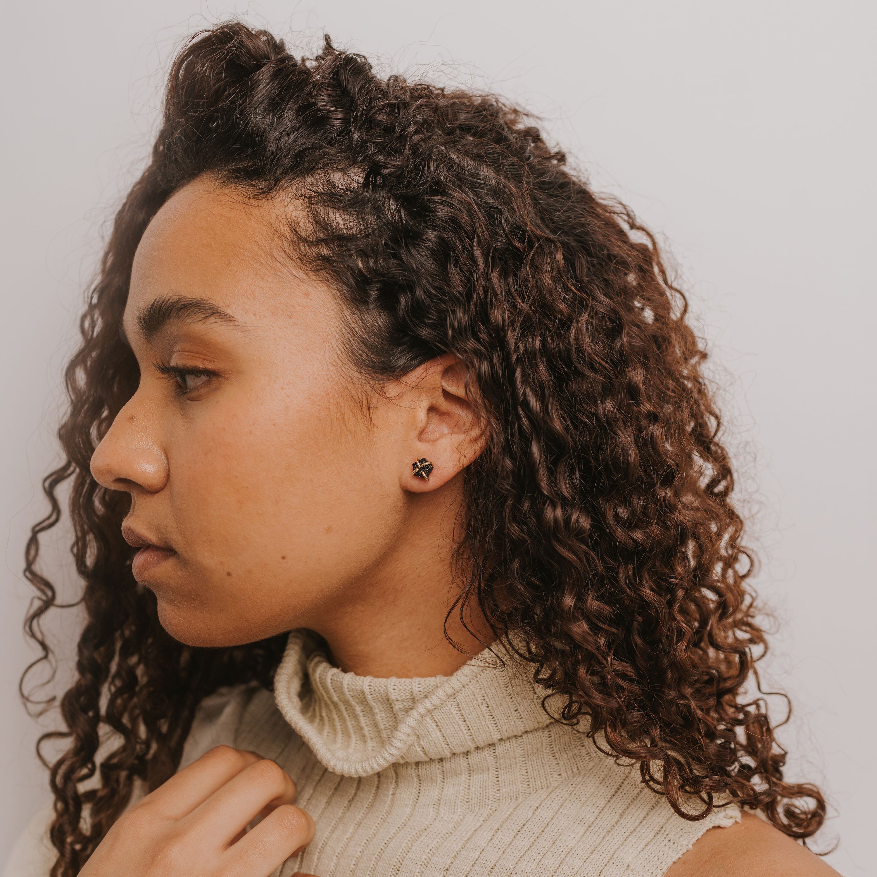 A woman with curly brown hair wears a cream turtleneck sweater and the Black Druzy Prong—a small, round stud earring with 18k gold plating—shown in profile against a plain background.