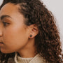 A person with curly hair wearing a cream sweater is shown in close-up, facing left, wearing a Black Druzy Prong stud earring. The background is plain and light-colored.