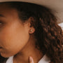 A close-up of a person with curly brown hair wearing a white hat and the Rose Gold Druzy Prong, a gold textured stud earring made from sterling silver, as their hand gently touches their neck.