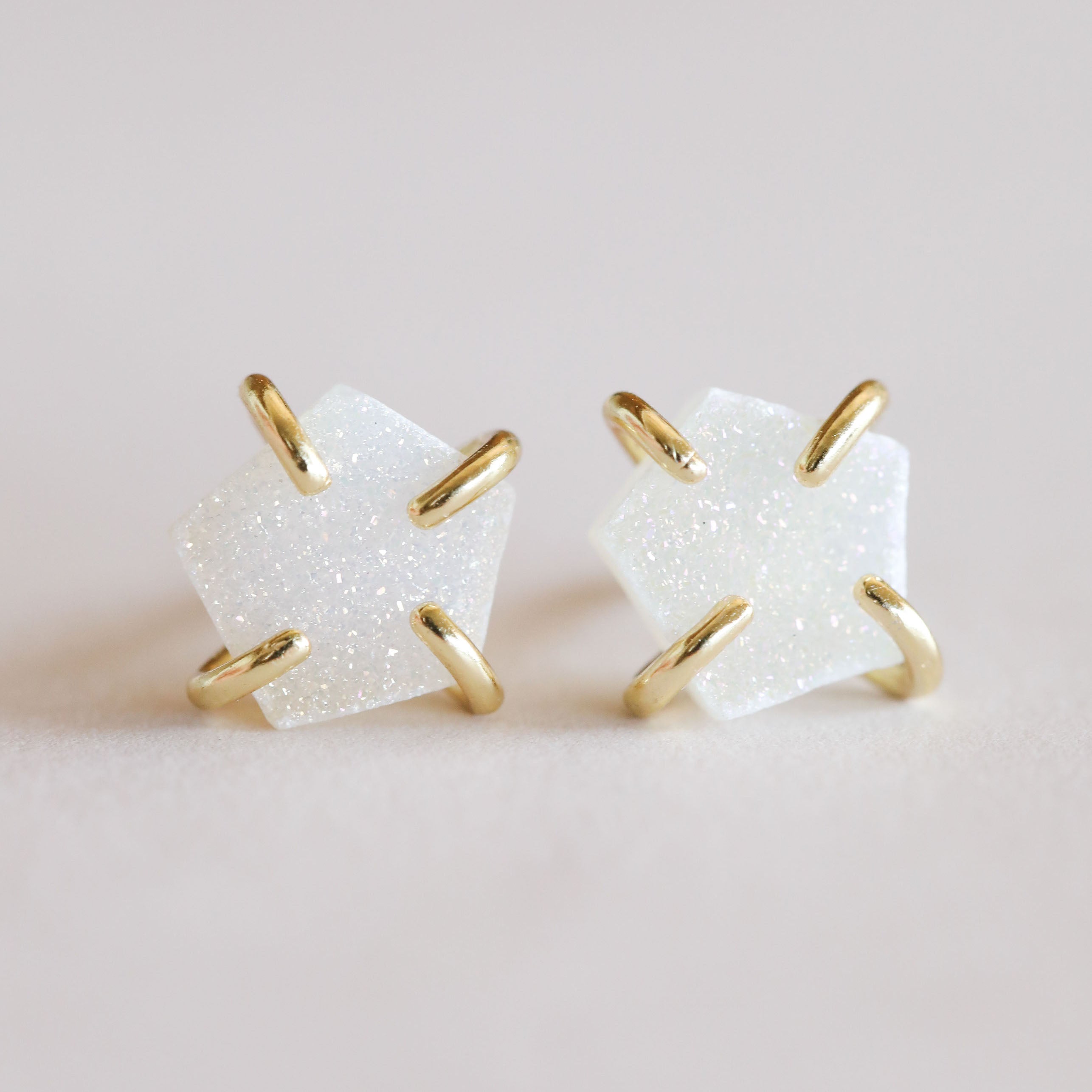 The White Druzy Prong features pentagon-shaped, glittery white natural druzy stone stud earrings set in 18k gold-plated prongs, shown on a soft beige background.