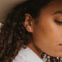 Close-up of a person with curly dark hair wearing a beige hat and the White Druzy Prong earring, crafted from sterling silver. The person is dressed in a white shirt and looking downward.