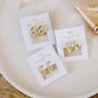 Three pairs of Gold Swirl Hoop gold-plated earrings are displayed on white "JAWKELLY wear good rules" cards, arranged on a cream dish. This set offers durable jewelry in classic hoop and textured designs.