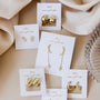 Five pairs of Gold Wide Hoop earrings with minimalist 18k gold-plated designs sit on white cards labeled "JAYCELLY wear good vibes." Hoops, studs, and dangles are arranged on a beige fabric background.