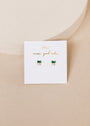 A white card with gold text "JaxKelly wear good vibes" showcases the Emerald Double Stud Stack—small rectangular emerald baguette stud earrings with gold accents and 18k gold plating—set against a soft beige background.
