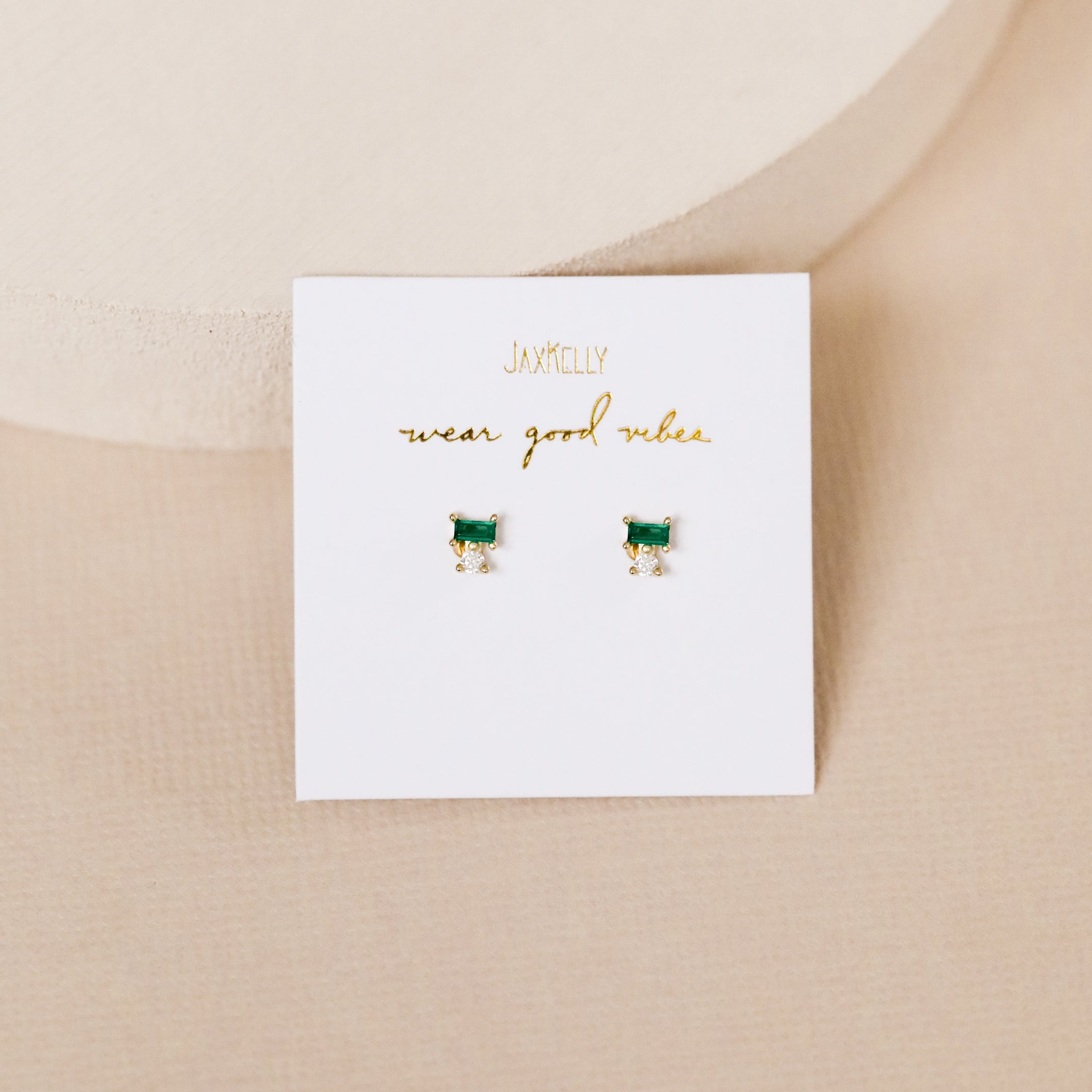 A white card with gold text "JaxKelly wear good vibes" showcases the Emerald Double Stud Stack—small rectangular emerald baguette stud earrings with gold accents and 18k gold plating—set against a soft beige background.