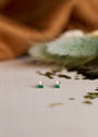 The Emerald Double Stud Stack features an emerald baguette and a CZ stone set in 18k gold plating, displayed on neutral fabric with a blurred brown and green background.