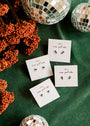 Four pairs of Emerald Double Stud Stack earrings with CZ stones and 18k gold plating on JANELLY cards, arranged on a green surface with orange berries and disco ball decorations.