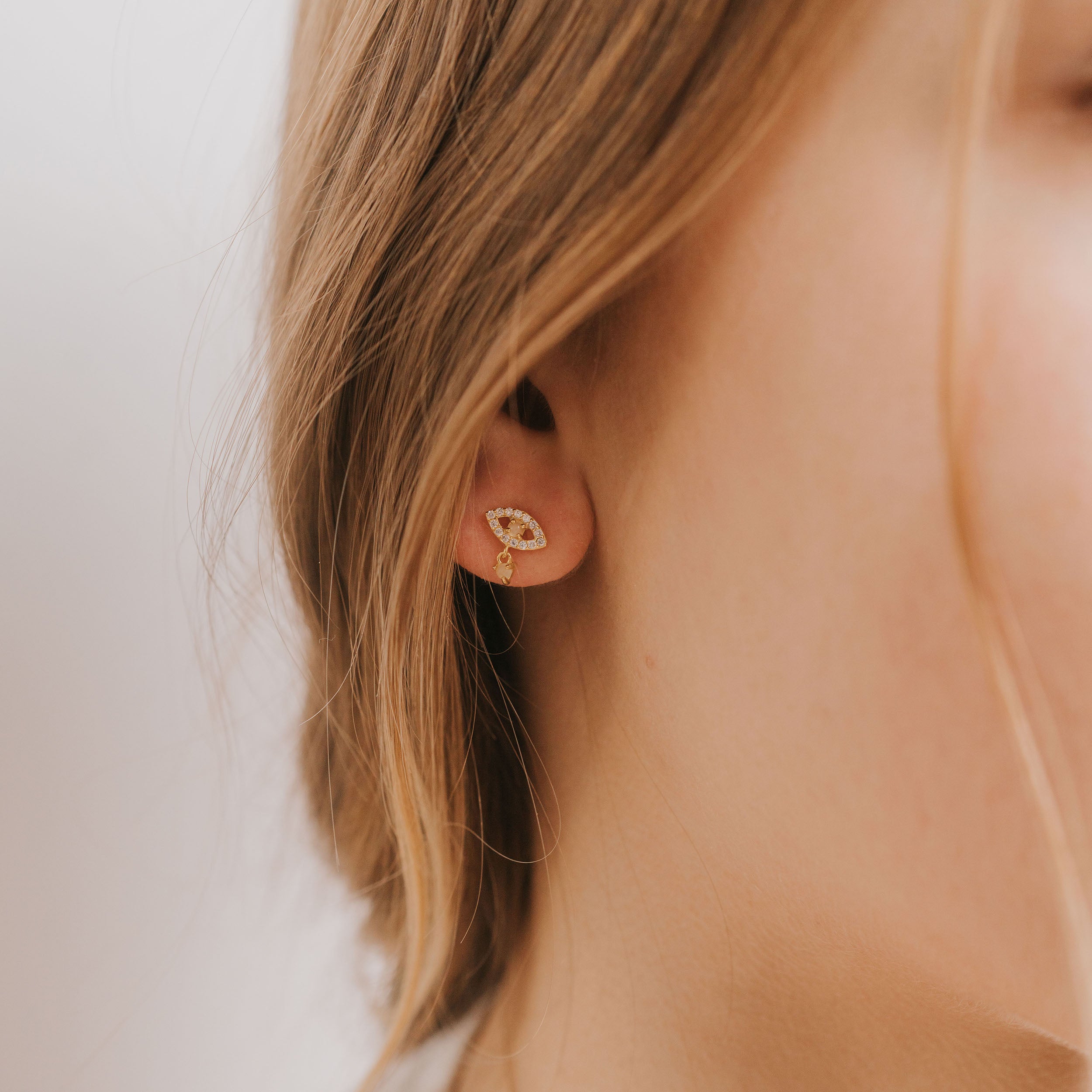 Close-up of a woman's ear with light brown hair, wearing the Mother of Pearl Evil Eye Drop earring—gold, teardrop-shaped, accented with CZ stones. A softly blurred background enhances its elegant detail.